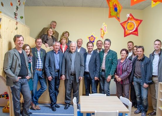 Eine Gruppe von Menschen in Anzügen steht lächelnd zusammen in einem Raum. Hinter ihnen ist eine Holzwand mit hängenden Sternen und Spielzeug dekoriert.