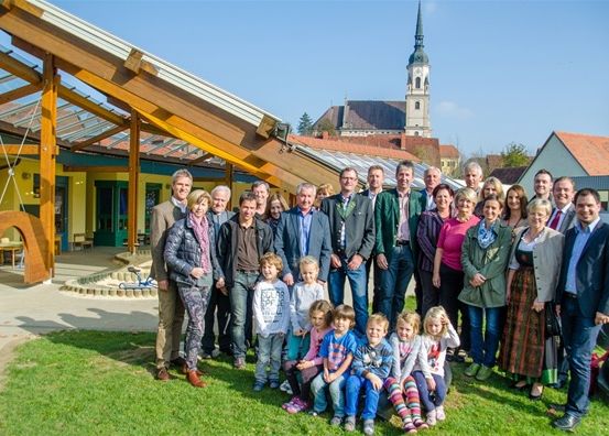 Eine Gruppe von Erwachsenen und Kindern posiert für ein Foto vor einem modernen Gebäude mit einem grünen Dach und einem Kirchturm im Hintergrund.