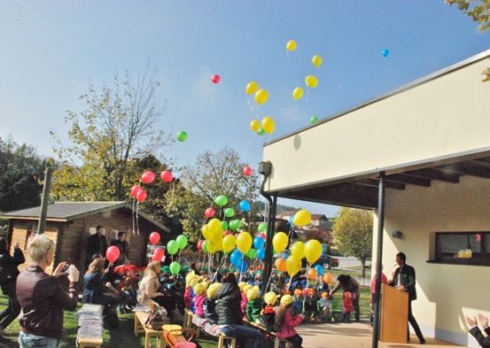 Eine Gruppe von Kindern und Erwachsenen versammelt sich im Freien in der Nähe eines Gebäudes, wobei Ballons in der Luft schweben. Ein Mann spricht an einem Rednerpult.
