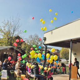 Eine Gruppe von Kindern und Erwachsenen versammelt sich im Freien in der Nähe eines Gebäudes, wobei Ballons in der Luft schweben. Ein Mann spricht an einem Rednerpult.