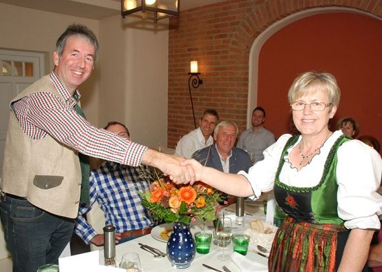 Eine Gruppe von Menschen versammelt sich in einem Esszimmer. Ein Mann und eine Frau in traditioneller Kleidung schütteln sich die Hände vor einem Tisch mit einem Blumenstrauß und Weingläsern.