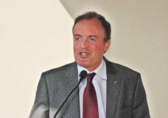 Ein Mann in formeller Kleidung, möglicherweise in ein Mikrofon sprechend, steht vor einer weißen Wand.