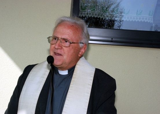 Ein älterer Mann mit einer weißen Stola und Brille spricht in ein Mikrofon. Hinter ihm ist eine Wand mit einem gerahmten Bild.