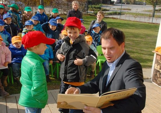Ein Mann liest Kindern in einem Park ein Buch vor. Die Kinder sitzen auf grünen Stühlen, und einige tragen Mützen.