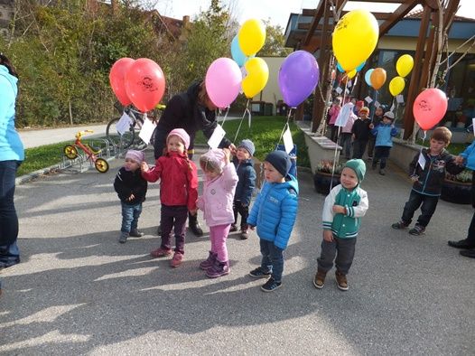Eine Gruppe von Kindern mit bunten Luftballons steht draußen in der Nähe eines Gebäudes, wobei eine Person dahinter einen Luftballon hält.
