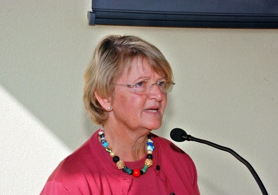 Eine ältere Frau mit kurzen blonden Haaren, die eine Brille und eine Halskette trägt, spricht in ein Mikrofon.