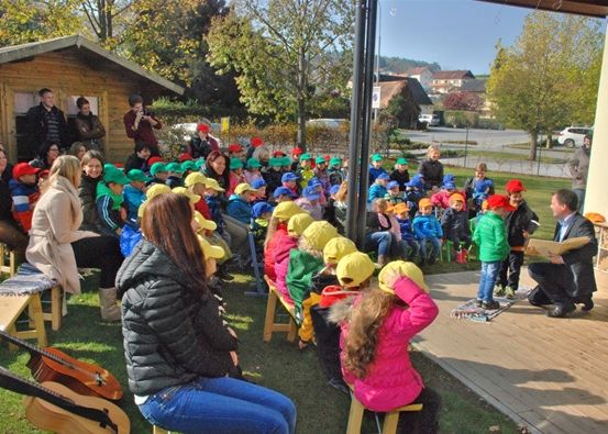 Eine Gruppe von Kindern in grünen Mützen sitzt auf Bänken in einer Freiluftumgebung. Ein Erwachsener liest aus einem Buch auf dem Boden.