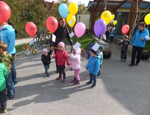 Kinder versammeln sich im Kreis und halten Luftballons. Sie sind von Erwachsenen umgeben, von denen einer ein Papier hält.