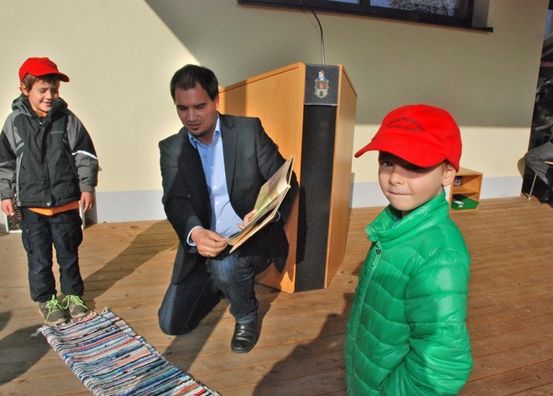 Ein Mann kniet und liest ein Buch vor zwei Jungen, einer in einer grünen Jacke, der andere in einer roten Mütze, in einer Innenumgebung. Hinter ihnen steht ein hölzerner Rednerpult mit einem Logo und einem Lautsprecher.