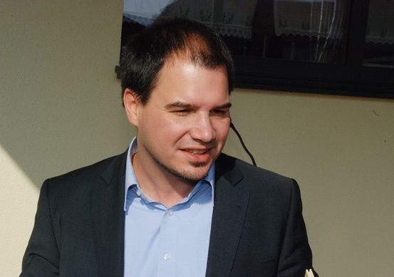 Ein Mann in einem blauen Hemd und einer schwarzen Anzugjacke lächelt in die Kamera. Er steht vor einer weißen Wand.