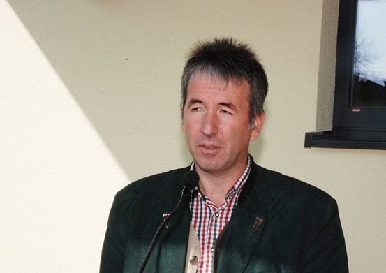 Ein Mann steht hinter einem Mikrofon, vor einer Wand mit einem Fenster im Hintergrund.