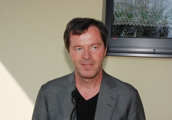 Ein Mann steht vor einem Mikrofon, lächelt und hat eine Wand und ein Bild hinter sich.