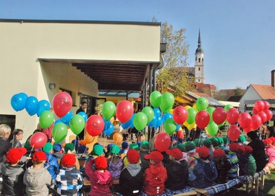 Eine Gruppe von Kindern und Erwachsenen versammelt sich im Freien, viele tragen rote Mützen. Sie halten bunte Luftballons, und einige stehen. Hinter ihnen befindet sich ein Gebäude mit Vordächern, und in der Ferne ist ein Turm zu sehen.