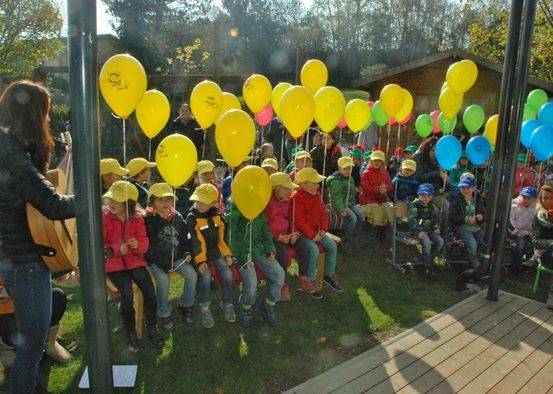 Eine Gruppe von Kindern in gelben Mützen sitzt auf einem Rasen und hält bunte Luftballons. In der Nähe spielt eine Frau Gitarre.