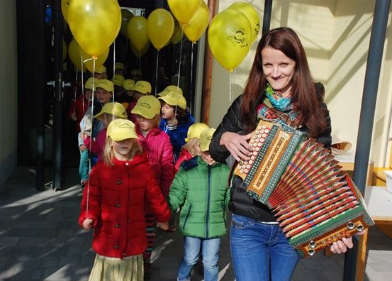 Eine Frau mit einem Akkordeon geht mit einer Gruppe von Kindern, einige halten Luftballons, in einem Flur.