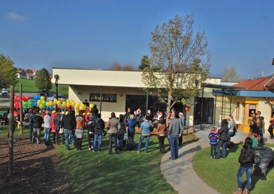 Eine Menschenmenge versammelt sich vor einem Gebäude, wahrscheinlich für eine Veranstaltung, mit Luftballons, geparkten Autos in der Nähe und Bäumen.