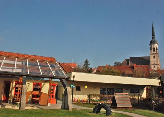 Ein Gebäude mit Glasdach, roten Fenstern und bunten Dekorationen davor, mit einem Kirchturm im Hintergrund.