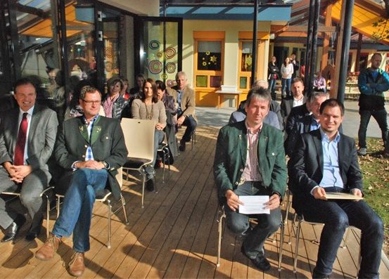 Eine Gruppe formell gekleideter Männer und Frauen sitzt auf einer Holzterrasse, wahrscheinlich bei einer Veranstaltung. Ein Mann hält ein Papier. Hinter ihnen sind Gebäude und Menschen zu sehen.