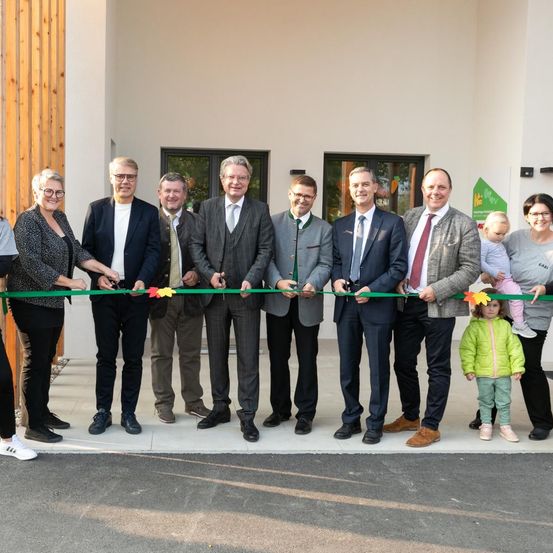 Mehrere Erwachsene und zwei Kinder stehen vor einem weißen Gebäude und schneiden ein grünes Band ab.