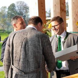 Drei Männer in Anzügen stehen vor einer Holzstruktur, einer hält eine Box. Ein Mann trägt eine grüne Krawatte, und ein anderer hat eine Brille. Bäume und ein Feld sind im Hintergrund.