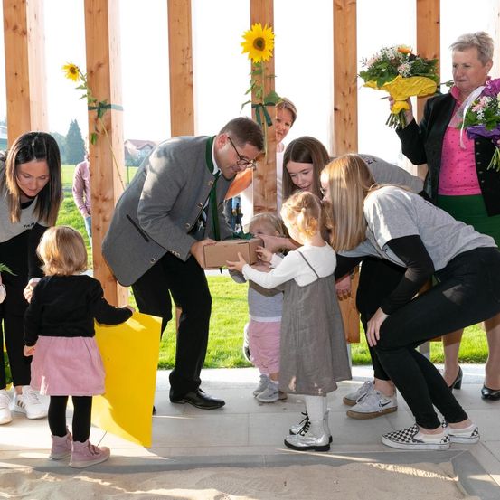 Eine Gruppe von Menschen steht im Kreis und hält Blumen und Geschenke. Ein Mann reicht einer kleinen Mädchen eine Box, während andere zusehen. Im Hintergrund befinden sich Holzstrukturen mit Sonnenblumen.