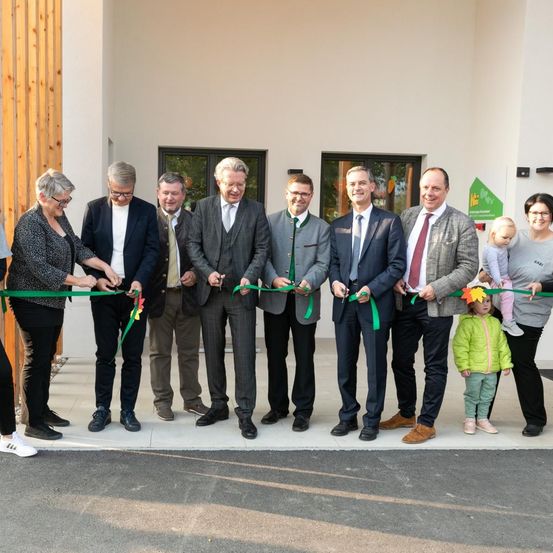 Eine Gruppe von Menschen, einschließlich Erwachsener und Kinder, steht für eine Eröffnungsfeier vor einem Gebäude.