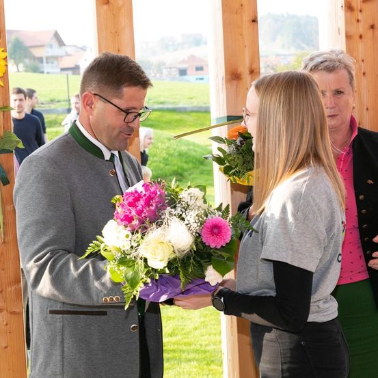 Ein Mann in einem grauen Mantel überreicht einer Frau in einem grauen Shirt einen Blumenstrauß unter einem hölzernen Pavillon.