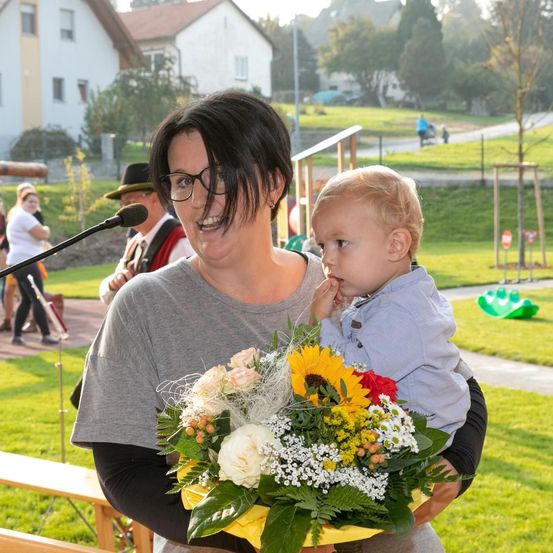 Eine lächelnde Frau mit Brille hält ein Baby und einen Blumenstrauß, während sie in einem Park mit einem Mikrofon in der Nähe steht.