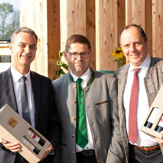 Drei Männer, in Anzügen gekleidet, stehen zusammen und halten Weinkartons in der Hand, einer trägt eine grüne Krawatte, der andere eine rote Krawatte, vor einer Holzwand.