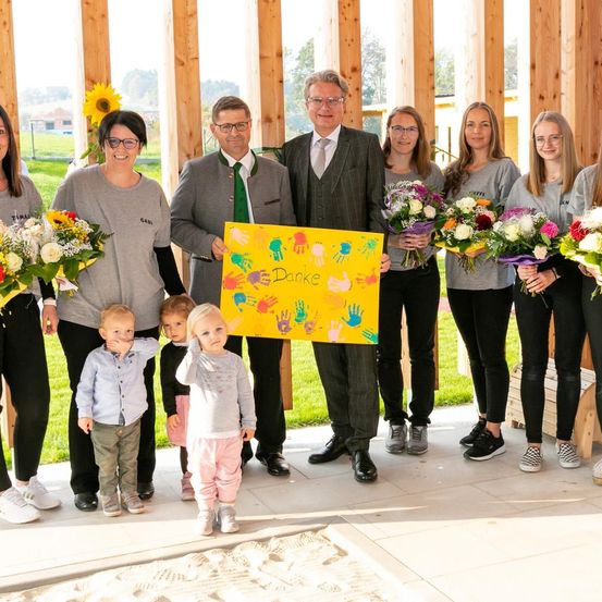 Eine Gruppe von Erwachsenen und Kindern steht in einer Reihe, lächelt und hält Blumen. Der Mann in der Mitte hält ein gelbes Schild mit dem Wort Danke. Dahinter steht eine Holzstruktur.