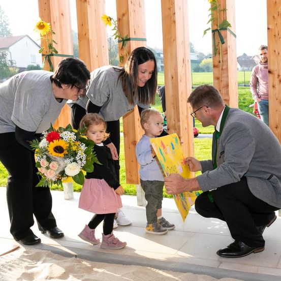 Ein Mann kniet und hält ein Schild für ein Kind zum Unterschreiben. Zwei Frauen und ein Kind mit Blumenstrauß schauen zu.