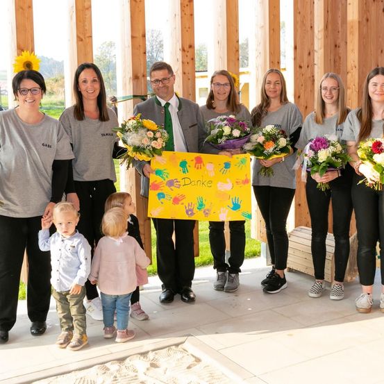 Eine Gruppe von Erwachsenen und Kindern steht in einer Reihe mit Blumen, einer hält ein Schild mit der Aufschrift Danke. Sie tragen alle Freizeitkleidung. Hinter ihnen steht eine Holzkonstruktion.