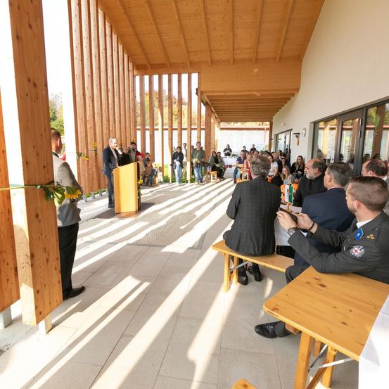 Eine formelle Veranstaltung findet in einem mit Holzpaneelen ausgestatteten Saal statt. Eine Person spricht an einem Rednerpult, und mehrere Personen sitzen auf Bänken und unterhalten sich. Der Boden ist mit Fliesen bedeckt, und Sonnenlicht erhellt den Raum.