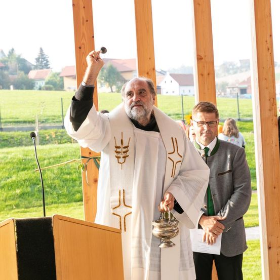 Ein Priester hält eine Rede im Freien, einen Kelch und eine kleine Glocke in der Hand. Ein anderer Mann in einem Anzug hält ein Papier. Der Priester steht vor einem Podium. Hinter ihnen sind ein Feld und Häuser sichtbar.
