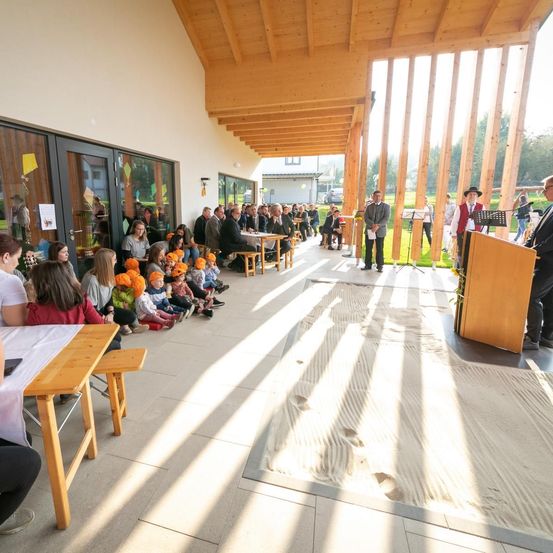 Eine Gruppe von Menschen sitzt auf Bänken und Stühlen in einem Saal. Eine Person steht an einem Podium und spricht die Zuhörer an. Der Saal hat ein Holzdach und Glaswände.