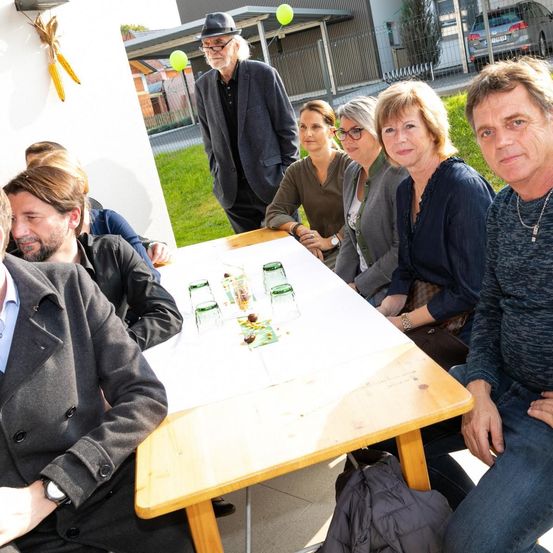 Eine Gruppe von Menschen sitzt um einen Tisch mit Gläsern und Essen darauf. Ein Mann steht hinter ihnen. Sie alle schauen auf etwas auf dem Tisch.