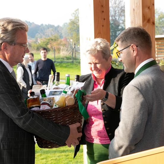 Ein Mann überreicht einer lächelnden Frau einen Korb mit Waren, während ein anderer Mann zusieht. Sie sind draußen mit einem grünen Rasen und Bäumen im Hintergrund.