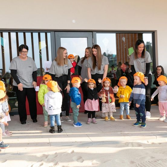 Eine Gruppe von Kindern und Erwachsenen steht auf einer sandigen Oberfläche. Sie tragen orangefarbene Mützen. Dahinter ist ein Gebäude mit Glastüren zu sehen.