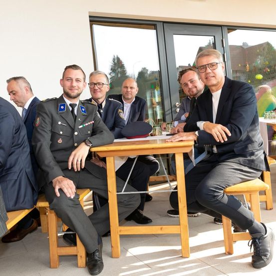 Sechs Männer in Anzügen und Militäruniformen sitzen an einem Holztisch, möglicherweise bei einer formellen Veranstaltung. Der Mann in Uniform trägt eine Uhr.