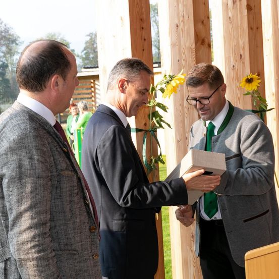 Drei Männer in Anzügen tauschen draußen eine Schachtel aus, einer trägt eine Brille und eine grüne Krawatte. Im Hintergrund sind Holzsäulen und Sonnenblumen zu sehen.