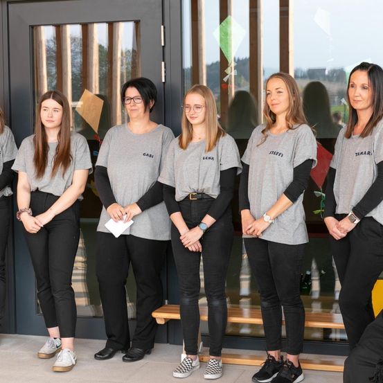 Fünf Frauen in übereinstimmenden grauen T-Shirts mit verschiedenen Namen stehen vor einem modernen Gebäude. Sie tragen alle schwarze Hosen und Turnschuhe, und einige von ihnen tragen Brillen. Die Glastür hat einen Holzrahmen.