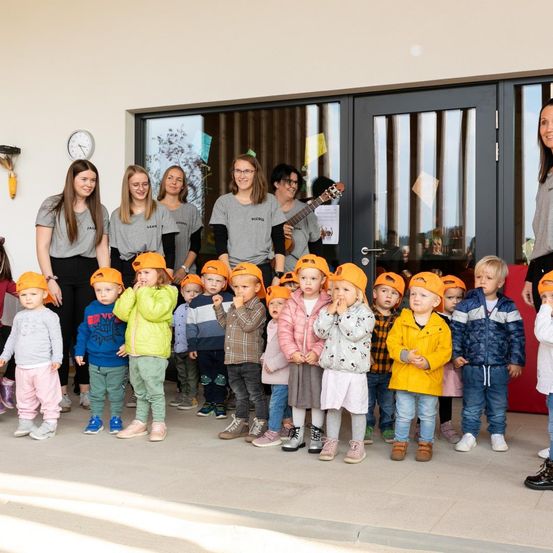 Eine Gruppe junger Kinder und Erwachsener steht gemeinsam vor einem Gebäude mit Glastüren und trägt orangefarbene Mützen.