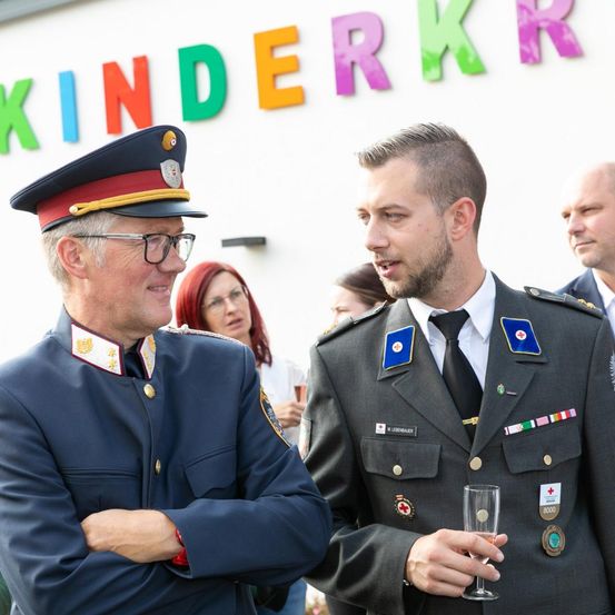 Zwei Männer in Uniformen stehen vor einem Gebäude. Einer hält ein Weinglas. Hinter ihnen stehen und reden Leute. Das Gebäude hat ein Schild, das 'KINDERKKR' steht.
