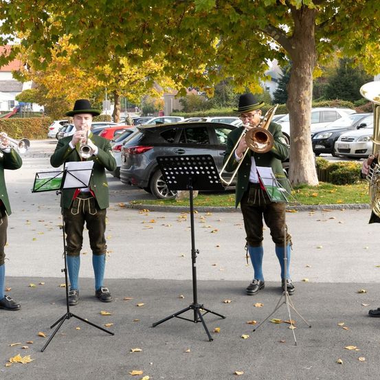 Vier Musiker in grünen Uniformen spielen Trompeten und Posaunen unter einem Baum auf einem Parkplatz. Blätter verteilen sich auf dem Boden, und mehrere Autos sind hinter ihnen geparkt.
