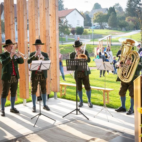 Vier Männer in traditioneller bayerischer Kleidung spielen Blechblasinstrumente auf einer Bühne mit Holzstrukturen und einer Bank im Hintergrund. Der Rasenbereich ist von Häusern umgeben.