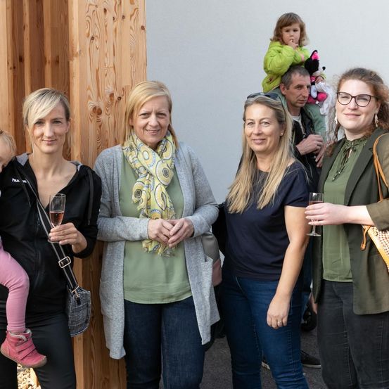 Fünf Frauen posieren für ein Foto. Die Frau auf der linken Seite hält ein Kind, und die Frau auf der rechten Seite hält ein Weinglas. Dahinter trägt ein Mann ein Kind auf seinen Schultern.