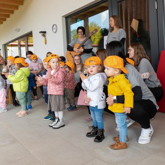 Mehrere Kinder stehen in einer Reihe und tragen orangefarbene Mützen. Sie schauen einer Frau zu, die Gitarre spielt. Dahinter stehen Erwachsene.