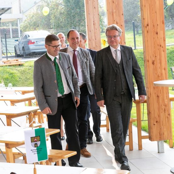 Mehrere Männer in Anzügen gehen auf einer Terrasse mit Holztischen und Bänken, vor einem mit einem Zaun umgebenen Grasbereich. Ein Mann trägt eine grüne Krawatte mit Logo. Dahinter ist ein Auto geparkt.