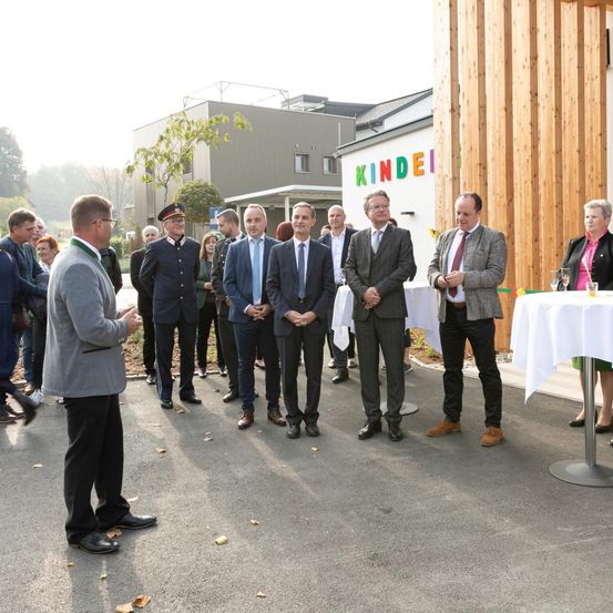 Mehrere Männer in Anzügen und Krawatten stehen in der Nähe eines Gebäudes mit dem Wort Kinder darauf. Sie scheinen an einer formellen Veranstaltung teilzunehmen.