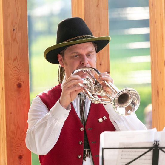 Ein Mann in einem roten Weste und schwarzem Hut spielt Trompete in einer Holzstruktur.